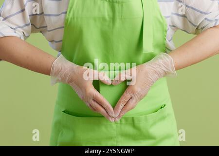 Nettoyage de printemps. Gros plan sur la femme moderne dans un tablier vert et des gants en caoutchouc sur fond vert avec une éponge de nettoyage montrant des mains en forme de cœur. Banque D'Images