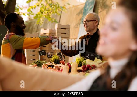 Objectif sélectif du fournisseur âgé accueillant le client afro-américain au stand greenmarket, offrant des produits biologiques frais. Des individus multiculturels souriants serrant la main au stand du marché alimentaire agricole. Banque D'Images