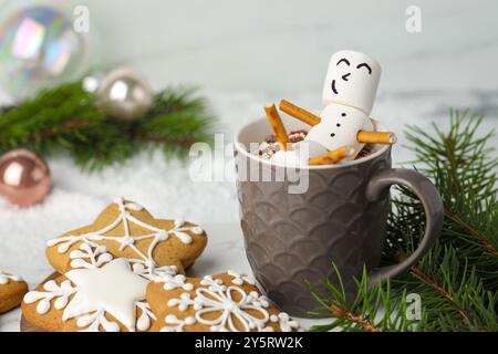 Joli bonhomme de neige guimauve dans une tasse de boisson chaude et des biscuits en pain d'épices sur une table blanche avec décor de Noël, gros plan Banque D'Images