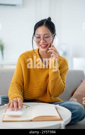 Une femme asiatique joyeuse dans un pull jaune confortable et des lunettes est assise sur un canapé à la maison, parlant au téléphone tout en faisant une liste dans un cahier. Banque D'Images