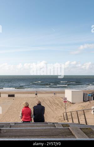 Egmond aan Zee, pays-Bas - 17 septembre 2024 : un couple vu de derrière se détendre sur des marches en bois vers une plage de sable tandis que d'autres se promènent le long de la plage Banque D'Images