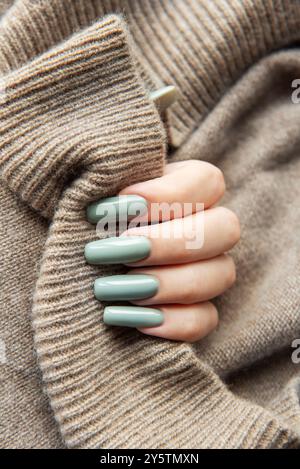 Une main féminine avec de longs ongles verts élégants tient doucement un pull beige doux, mettant en valeur une esthétique confortable et à la mode parfaite pour l'automne. Banque D'Images