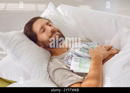 Homme endormi souriant couché dans son lit avec les yeux fermés, embrassant beaucoup de billets de banque en dollars à la maison Banque D'Images