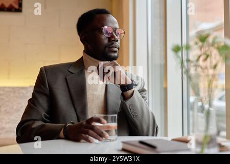 Homme portant des lunettes de soleil assis à la table du café, tenant un verre d'eau et regardant à l'extérieur de la fenêtre, créant une atmosphère pensive avec un cahier sur la table Banque D'Images