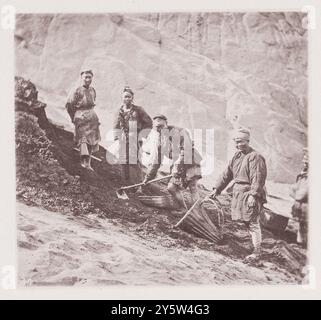 Photo vintage de mineurs de charbon chinois. Badong, province de Hubei. Illustrations de la Chine Qing, 1875 il y a un certain nombre de mines de charbon près de Badong, et dans les roches où se trouvent les lits de charbon les strates de calcaire ont été projetées du ruisseau dans des murs presque perpendiculaires. Les mineurs, dont un groupe est représenté sur cette photo, travaillent tous les jours de sept heures à quatre heures, et leur salaire moyen est de 300 espèces par jour, soit environ sept shillings par homme par semaine. Ils utilisent une petite lampe à huile fixée à la tête, semblable à celle que nos mineurs utilisaient avant l'invention de la lampe Davy. Banque D'Images