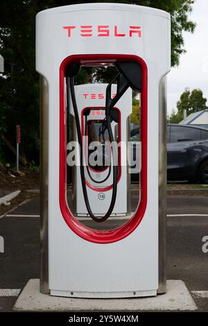 Borne de recharge Tesla blanche et rouge dans un parking de banlieue, avec véhicule électrique partiellement obscurci en arrière-plan Banque D'Images