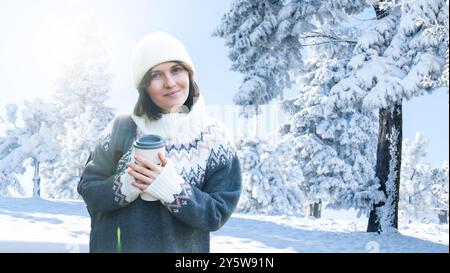 Femme vêtue d'un pull d'hiver tient une tasse avec une boisson chaude dans ses mains. Femme sur le fond de la forêt d'hiver.. Banque D'Images