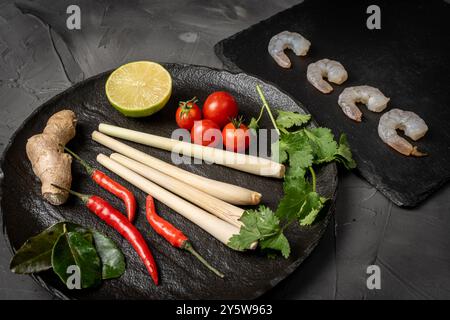 Ingrédients pour faire Tom Yum Kung. Citronnelle, piment, gingembre, galanga, citron vert, coriandre, tomates cerises et crevettes crues sur une assiette noire à nouveau Banque D'Images