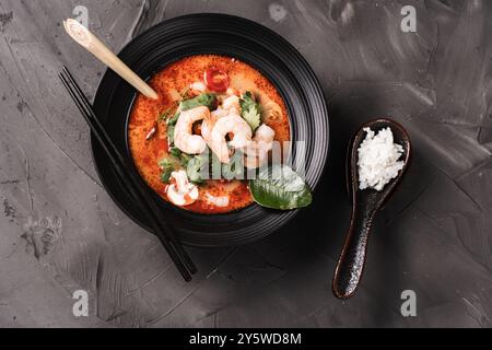 Tom Yam Kung soupe thaïlandaise épicée avec crevettes, citronnelle et galanga dans un bol noir sur fond sombre et une cuillère avec du riz. vue de dessus. Haute qualité p Banque D'Images