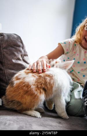 Un jeune enfant caresse doucement un lapin moelleux sur le canapé tout en profitant d'un après-midi paisible à la maison. L'espace de vie confortable est lumineux et accueillant. Banque D'Images