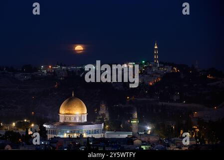 Une pleine lune orange s'élève bas à l'horizon au-dessus du mont Scopus, du dôme du Rocher et de la vieille ville de Jérusalem, en Israël. Banque D'Images