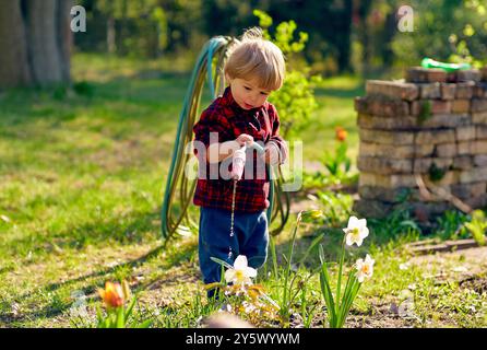 Enfant en bas âge dans une chemise à carreaux rouge jouant avec un tuyau d'arrosage parmi les fleurs de printemps dans une cour ensoleillée. Banque D'Images