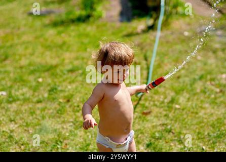 Enfant en bas âge jouant avec un tuyau d'arrosage par une journée ensoleillée, pulvérisant de l'eau à partir de la buse pendant qu'ils marchent à travers l'herbe. Banque D'Images