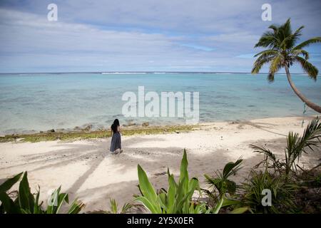 Scène de plage tropicale sereine avec une seule personne debout près du bord de l'eau sous un palmier, regardant l'océan, Titikaveka, Rarotonga, Îles Cook Banque D'Images