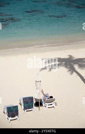 Femme lisant un e-book à la plage, chaises longues blanches et un parasol fermé, un palmier jette une ombre près de l'eau turquoise claire, Titikaveka, Rarotonga, Îles Cook Banque D'Images