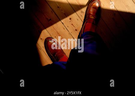 La lumière du soleil projette des ombres autour des chaussures brunes brillantes d'une personne sur un plancher en bois. Banque D'Images