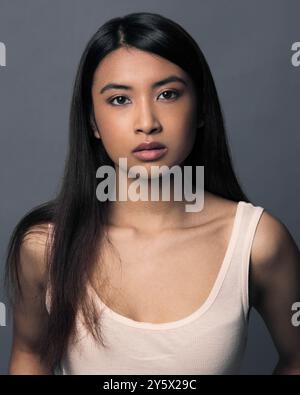 Portrait en gros plan d'une jeune femme aux cheveux longs et à l'expression neutre sur fond gris. Banque D'Images