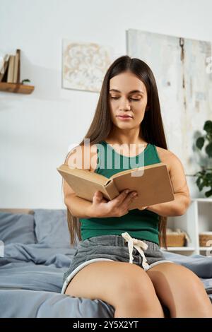 Une jeune femme brune à la petite stature se détend sur son lit, entièrement immergée dans un livre lors d'un après-midi confortable à la maison. Banque D'Images