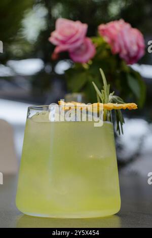 Un cocktail vert rafraîchissant garni d'une tranche d'ananas séchés et de romarin, avec des roses roses en arrière-plan. Banque D'Images