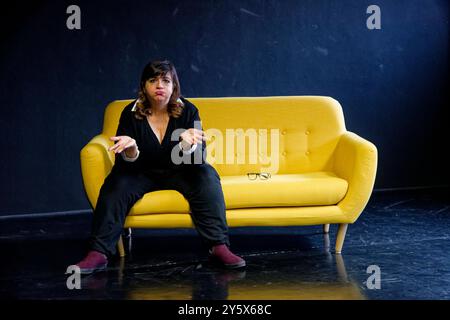 Femme dans une tenue noire assise sur un canapé jaune sur un fond noir avec une paire de lunettes à côté d'elle. Banque D'Images