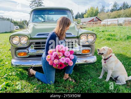 Une femme s'agenouille à côté d'un camion vintage tenant un bouquet de fleurs, avec un retriever doré assis à côté d'elle dans un champ herbeux. Banque D'Images