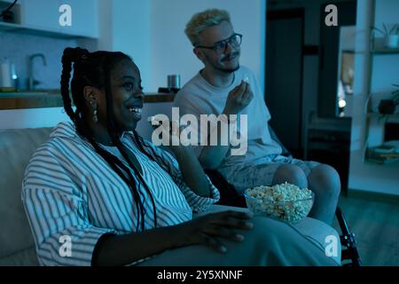 Heureux couple assis sur un canapé appréciant pop-corn tout en regardant un film dans un salon confortable. Souriant et appréciant les loisirs et la convivialité avec des en-cas Banque D'Images