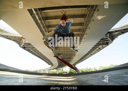 Skateboarder effectuant un tour sous un grand pont supérieur montrant l'habileté et l'agilité en plein air avec le skateboard retournant la tête en bas avec un fond structuré en métal et béton Banque D'Images