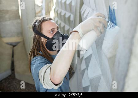 Artiste graffiti peignant une murale détaillée sur un mur extérieur, portant un masque respiratoire et des gants de protection, en se concentrant sur des motifs et des textures uniques dans le processus de création murale Banque D'Images