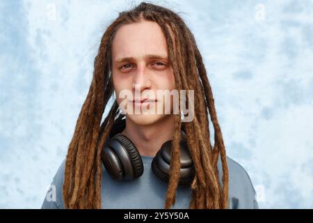 Portrait d'un jeune homme avec de longs dreadlocks portant de grands écouteurs autour du cou, debout devant un fond bleu pâle, exsudant la confiance et un comportement détendu Banque D'Images