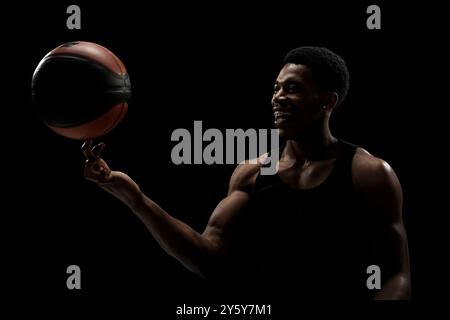 Joueur de basket-ball tournant une balle sur fond noir. Bonne silhouette d'homme afro-américain souriant. Banque D'Images