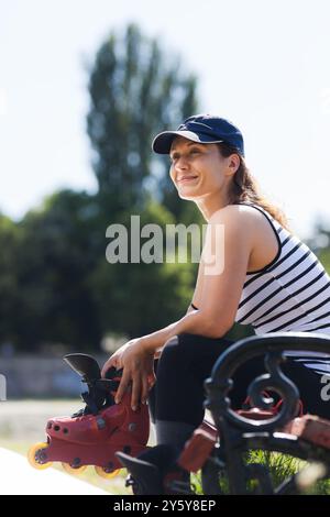 Une jeune femme souriante profite d'une journée ensoleillée dans le parc, assise sur un banc avec ses patins à roues alignées à la main, entourée par la nature. Banque D'Images