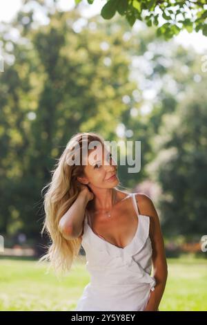 Une femme blonde gracieuse dans une robe blanche élégante pose naturellement dans un parc verdoyant, avec la lumière du soleil filtrant à travers les arbres. Banque D'Images