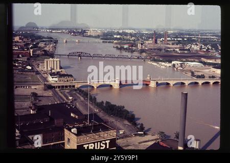 Une vue aérienne de Toledo, Ohio, vers les années 1970, montrant la rivière Maumee et ses ponts emblématiques. Le centre-ville historique est visible au premier plan, avec des complexes industriels et des lignes de chemin de fer qui s'étendent vers l'horizon, illustrant le patrimoine industriel de la ville au milieu du XXe siècle. Banque D'Images