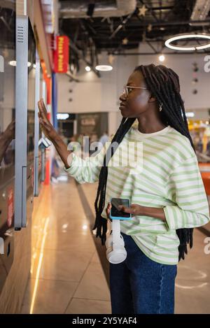 Une femme afro-américaine interagit avec un kiosque libre-service dans un environnement de vente au détail, tenant un smartphone et des écouteurs, concentrée sur sa tâche. Banque D'Images