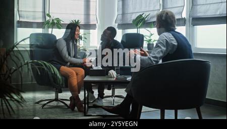 Couple multiethnique en séance de conseil avec thérapeute. Psychologue mature parle à la jeune famille. Homme afro-américain et femme asiatique assis sur des chaises tenant les mains après la querelle sur la thérapie de couple. Banque D'Images