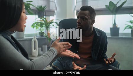 Le petit ami afro-américain en colère perd son sang-froid, commence à se battre et crie à sa petite amie pendant la thérapie de couple. Jeune couple multiethnique ayant un conflit en séance de conseil avec psychologue. Banque D'Images
