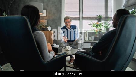 Vue arrière d'un couple multiethnique assis sur des chaises, discutant des problèmes relationnels, trouvant une solution et un compromis sur la séance de thérapie de couple. Thérapeute mature écoute la jeune famille, prend des notes. Banque D'Images