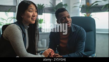 Heureux couple multiethnique en séance de conseil avec thérapeute. Homme afro-américain et femme asiatique assis sur des chaises tenant les mains après la querelle sur la thérapie de couple. Psychologue parle à la jeune famille. Banque D'Images
