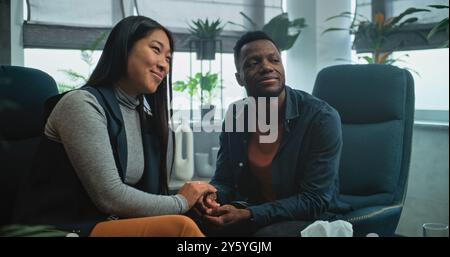 Heureux couple multiethnique en séance de conseil avec thérapeute. Homme afro-américain et femme asiatique assis sur des chaises tenant les mains après la querelle sur la thérapie de couple. Psychologue parle à la jeune famille. Banque D'Images