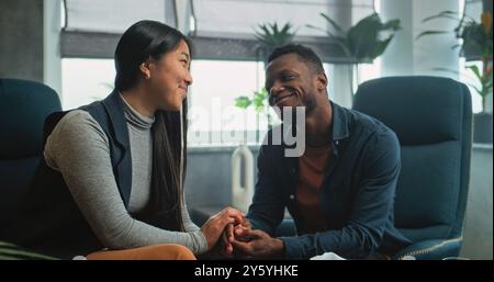 Heureux couple multiethnique en séance de conseil avec thérapeute. Homme afro-américain et femme asiatique assis sur des chaises tenant les mains après la querelle sur la thérapie de couple. Psychologue parle à la jeune famille. Banque D'Images