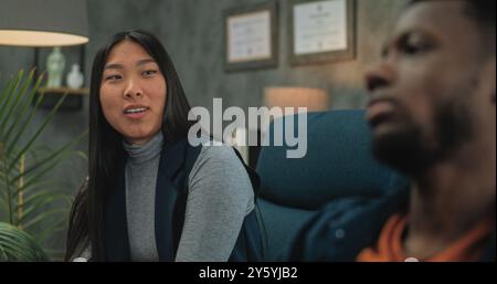Jeune couple multiethnique en séance de conseil avec psychologue. Une jeune femme asiatique parle de conflit relationnel. Au premier plan, malheureux petit ami afro-américain non focalisé. Thérapie de couple. Banque D'Images