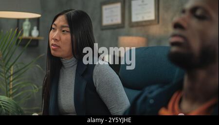 Jeune couple multiethnique en séance de conseil avec psychologue. Une jeune femme asiatique parle de conflit relationnel. Au premier plan, malheureux petit ami afro-américain non focalisé. Thérapie de couple. Banque D'Images