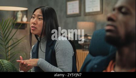 Jeune couple multiethnique en séance de conseil avec psychologue. Une jeune femme asiatique parle de conflit relationnel. Au premier plan, malheureux petit ami afro-américain non focalisé. Thérapie de couple. Banque D'Images