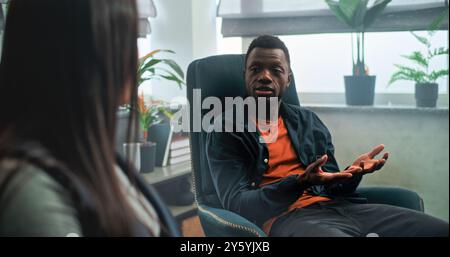 Le petit ami afro-américain en colère perd son sang-froid, commence à se battre et crie à sa petite amie pendant la thérapie de couple. Jeune couple multiethnique ayant un conflit en séance de conseil avec psychologue. Banque D'Images