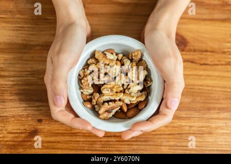 Gros plan des mains d'une femme tenant un bol en céramique blanche rempli de noix mélangées, y compris des noix et des amandes. Snack sain sur fond de table en bois, Banque D'Images