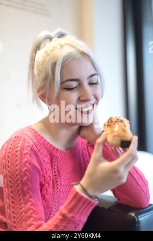 Une jeune femme à l'expression joyeuse profite d'une pâtisserie, vêtue d'un pull rouge vif, dans un cadre intérieur confortable avec une lumière naturelle qui l'illumine Banque D'Images