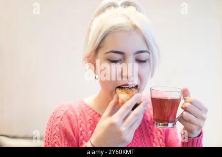 Une jeune femme savoure une pâtisserie tout en tenant une boisson chaude, son expression de délice, dans un cadre intérieur tamisé, mettant en valeur une atmosphère détendue Banque D'Images