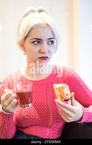 Une jeune femme savoure une pâtisserie tout en tenant une boisson chaude, son expression de délice, dans un cadre intérieur tamisé, mettant en valeur une atmosphère détendue Banque D'Images