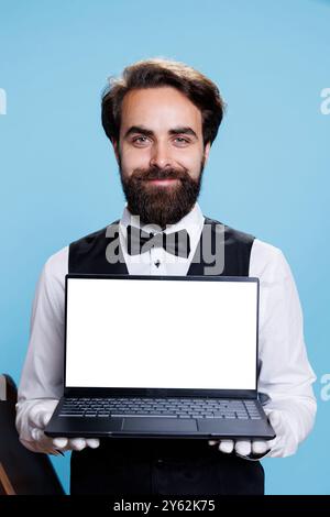 Jeune chasseur tient l'affichage blanc sur ordinateur portable dans le studio tandis qu'il porte costume formel et cravate à côté des bagages. Portier chic travaillant à l'hôtel de luxe et montre un modèle d'écran vierge. Banque D'Images