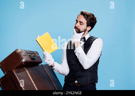 Wise Bellhop lit livre de littérature à côté de pile de bagages sur fond bleu, appréciant l'histoire de roman ou de fiction. Jeune homme avec salle de conférence lisant de la poésie pour la connaissance, portier intelligent. Banque D'Images